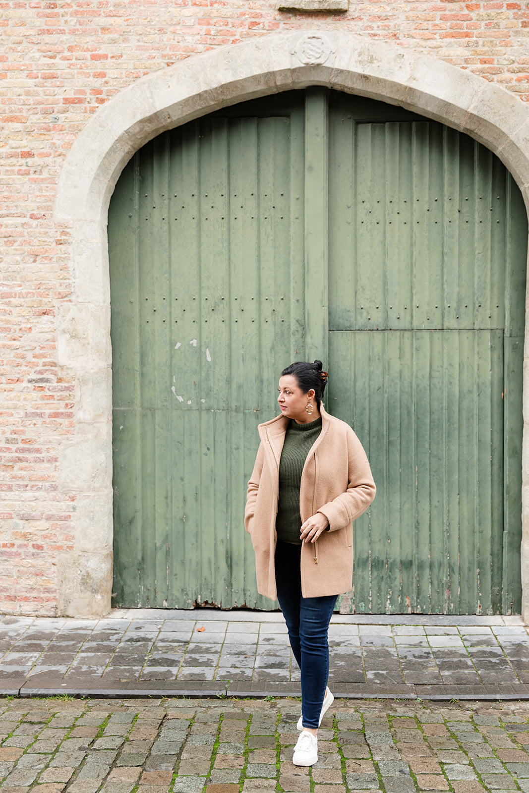 Woman in a beige coat standing in front of a large green door on a brick building.